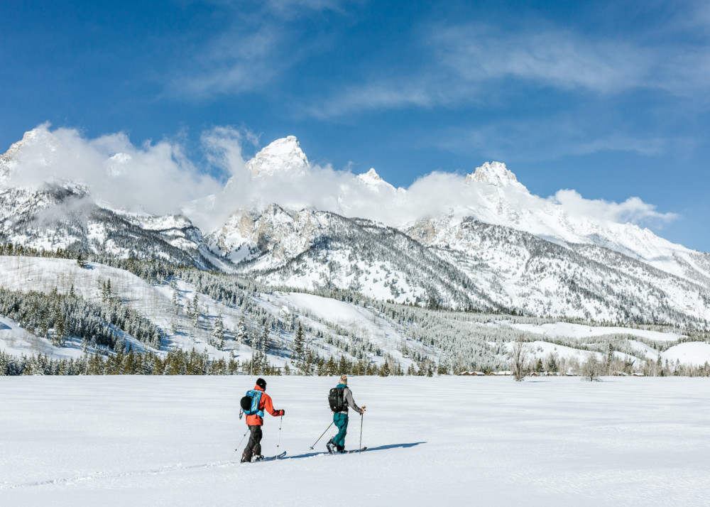 Two skiers tour across a flat field at the base of the Teton mountains.