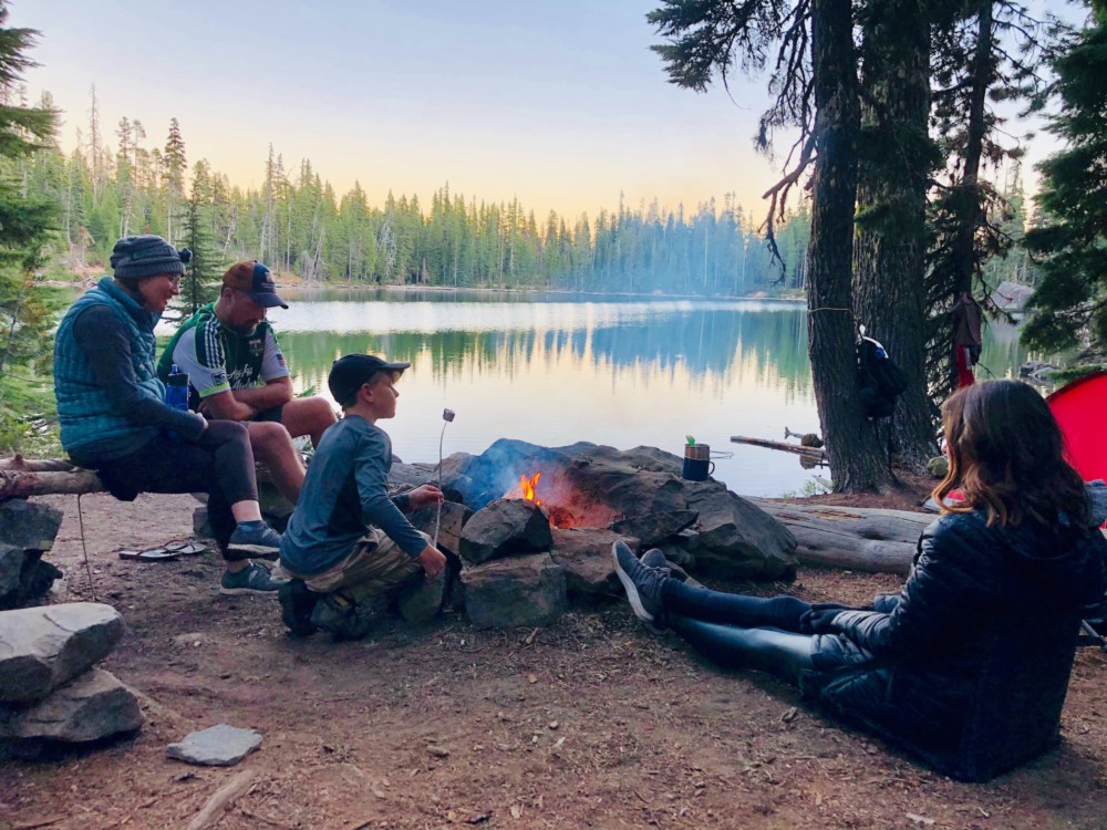 A family sits by a fire lakeside during a camping trip.