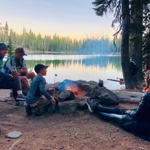 A family sits by a fire lakeside during a camping trip.