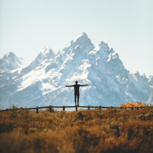 The silhouette of a man standing on a fence in front of the snowy Tetons on a fall day.