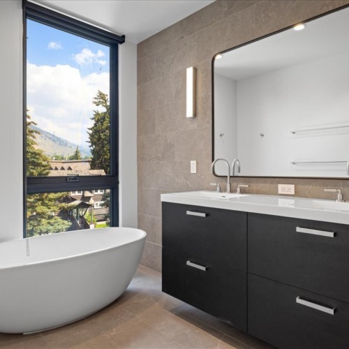 A modern bathroom with a black double vanity, beige tiled wall, large white bathtub, and mountain views.